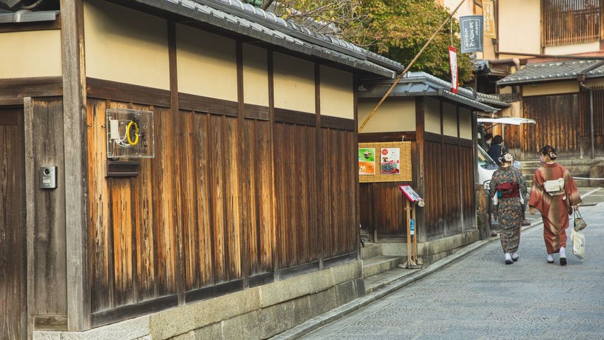 Back view of unrecognizable local Japanese ladies in traditional kimonos walking on paved street near typical aged wooden houses in Kyoto